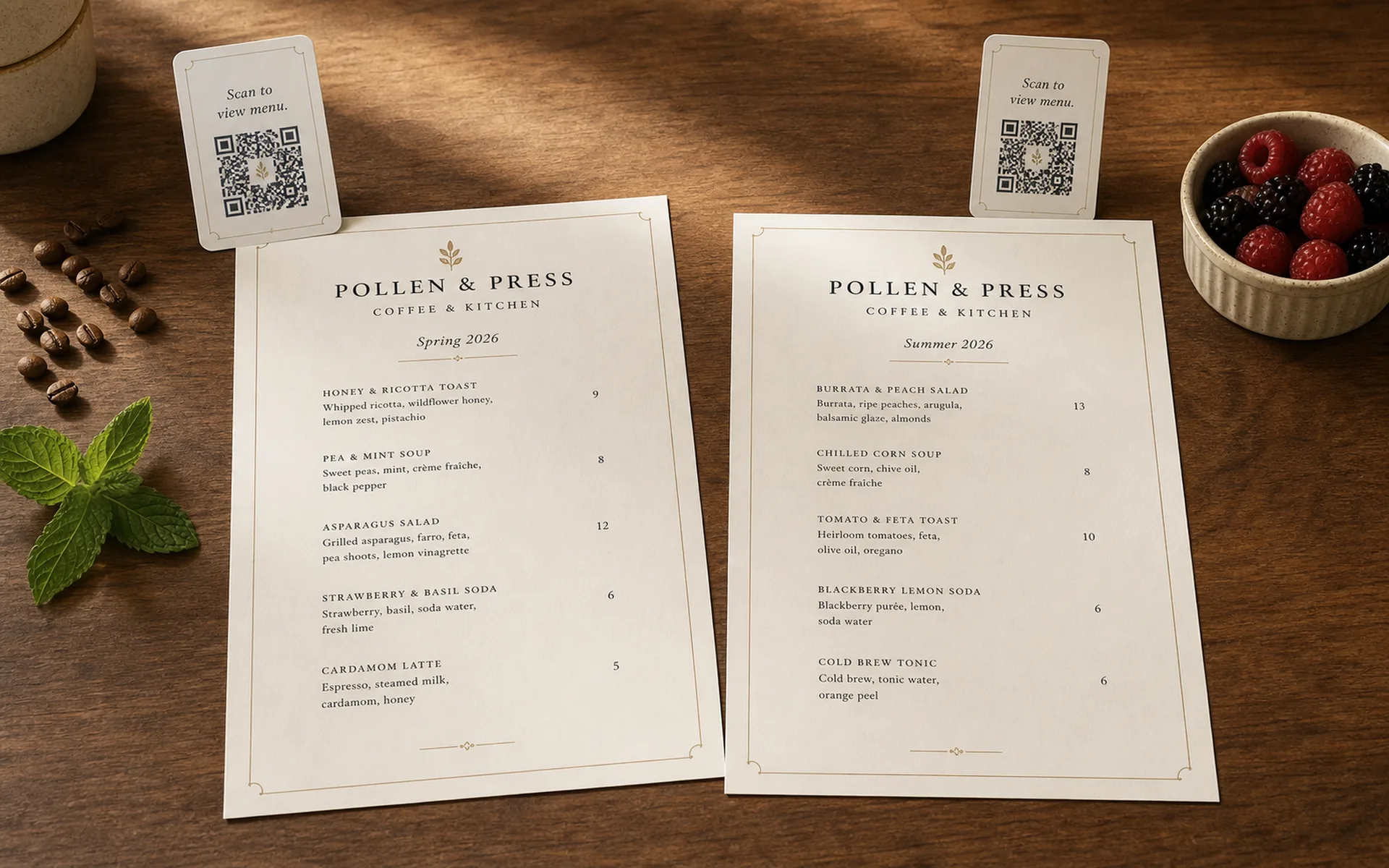 Two printed Pollen & Press cafe menu editions side by side on warm walnut — Spring 2026 and Summer 2026 — each with its own QR card and seasonal accents